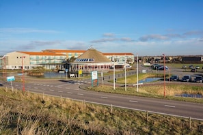 A building with a large central atrium, a parking lot, and a road with a pedestrian crossing. - Holiday home Strandperle 174 (Julianadorp)