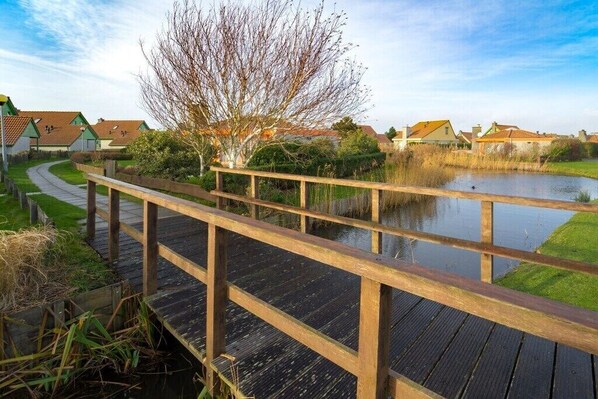 A wooden bridge over a calm water body, surrounded by greenery and residential buildings. - Holiday home Strandperle 174 (Julianadorp)