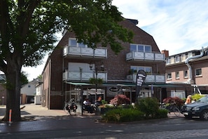 Ein zweigeschossiges Gebäude mit einem Café namens 'Le cafe' im Erdgeschoss, mit Außenbestuhlung und einem Schild für 'Brot & Butten'.