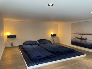 Een slaapkamer met een bed, twee wandlampen om te lezen, een schilderij van een boot op het water en een houten vloer.