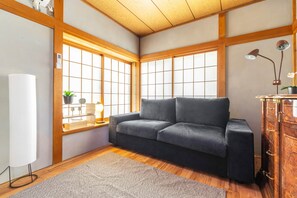 A room with a grey sofa, wooden beams, a window with a grid pattern, a desk with a lamp, and a cabinet with drawers.