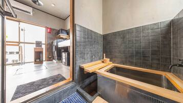 A modern bathroom with a wooden bathtub, a black tiled wall, and a visible laundry area in the background.