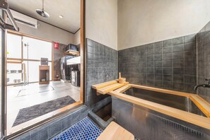 A modern bathroom with a wooden bathtub, a black tiled wall, and a visible laundry area in the background. - Skytree Next Door | Modern Ryokan in Tokyo (Katsushika City)