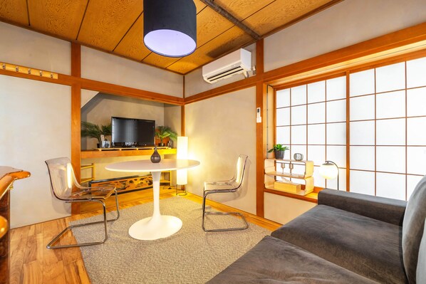 A modern living room with a sofa, a dining table with chairs, a TV, and a large window with a view.