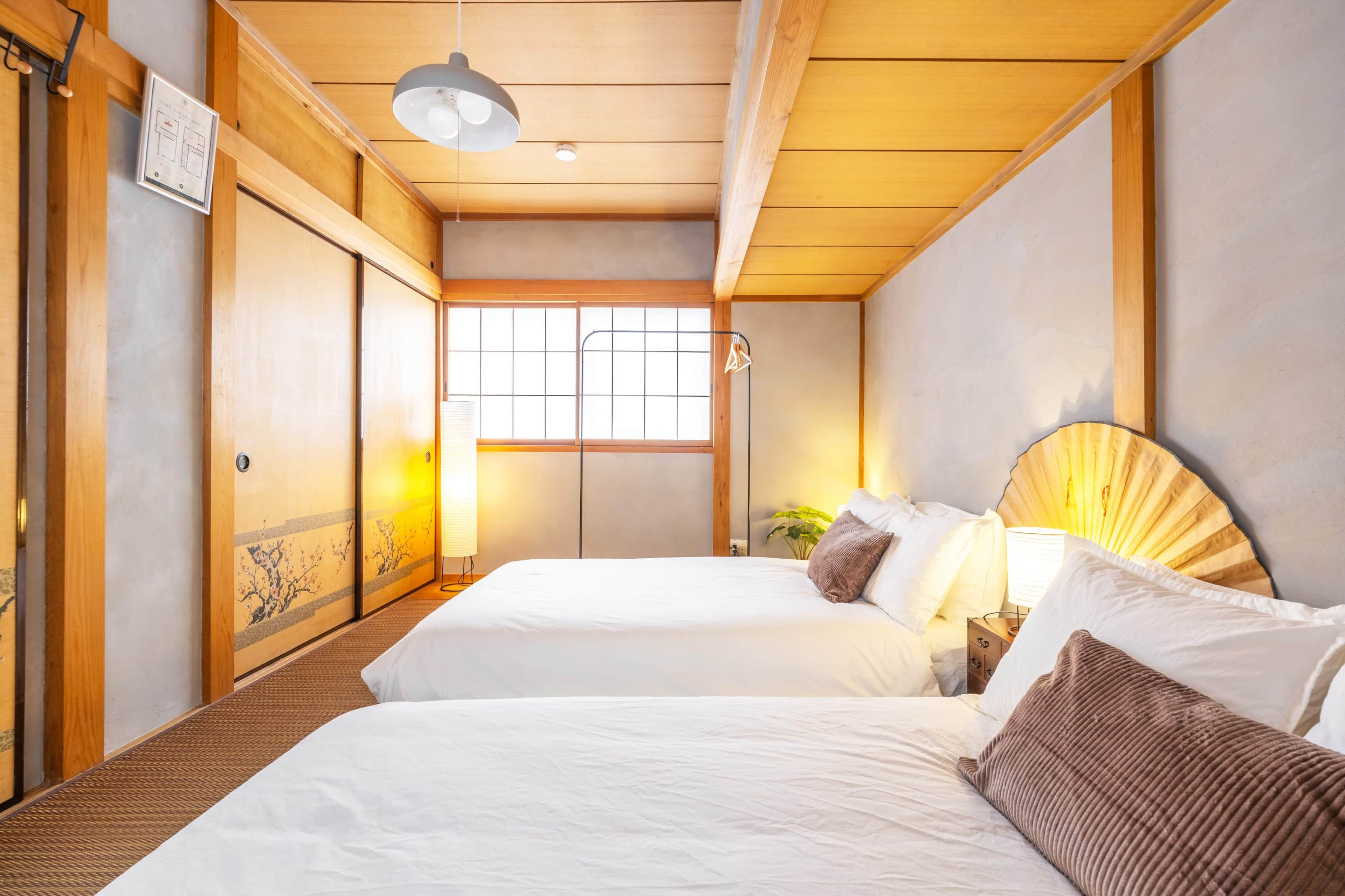 A hotel room with two beds, a traditional Japanese sliding door, a window with a view, and a bedside table with a lamp.