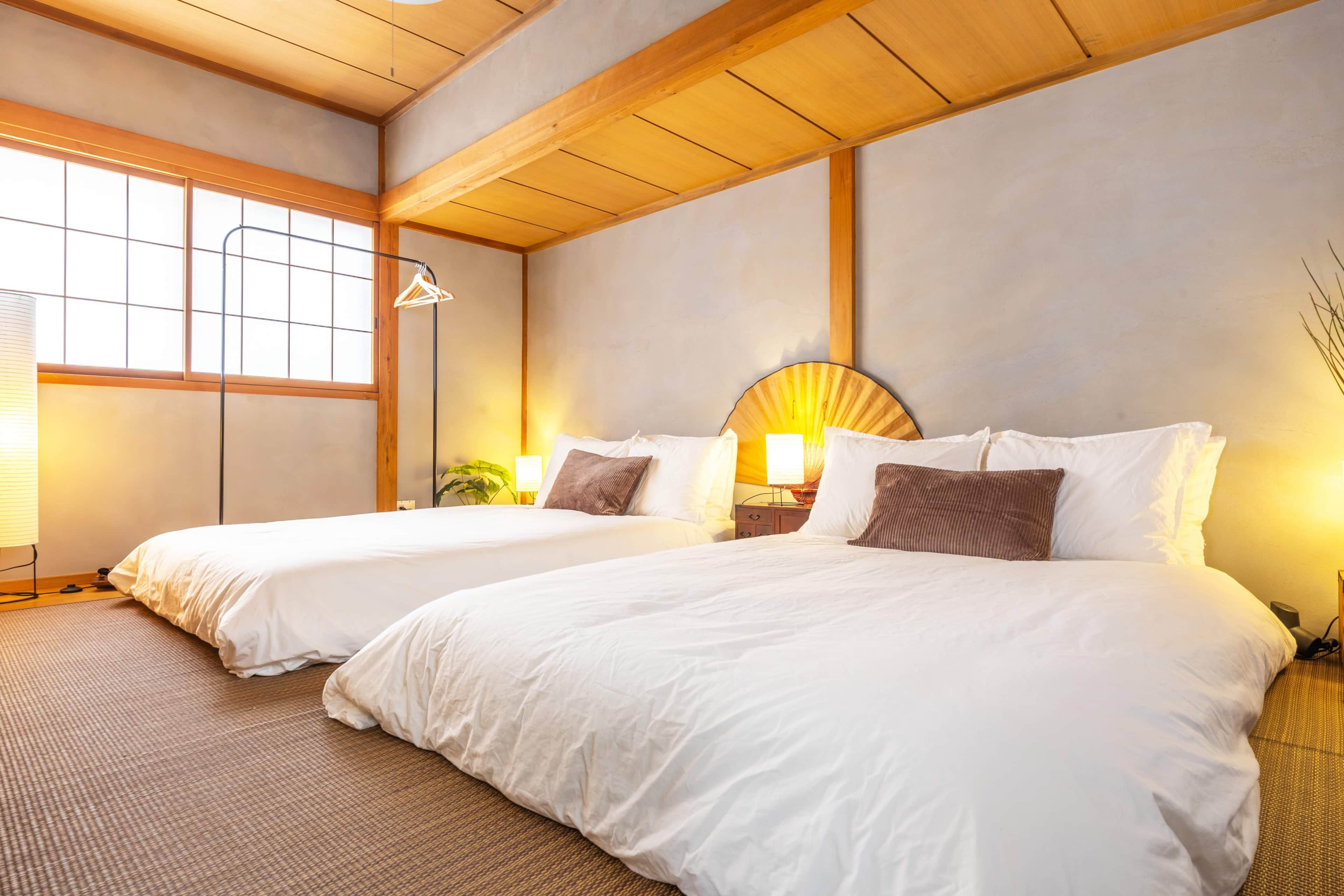 Quarto de hotel com duas camas, uma janela grande, um abajur e um ventilador fixado na parede.