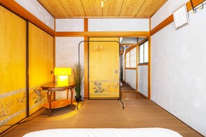 A traditional Japanese room with tatami flooring, a shoji screen door, a wooden table with a lamp, and a view of a hallway.