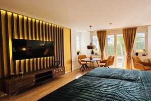 Ein Hotelzimmer mit einem großen Flachbildfernseher, einem Bett mit dunkelgrüner Decke, einer Holzunterhaltungsecke, einem kleinen runden Tisch mit Stühlen und einem Fenster mit Vorhängen.