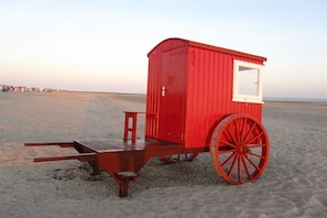 A red wooden structure with a window and a large wheel, situated on a sandy beach. - No. 8 in the Kiebitz House (Borkum)