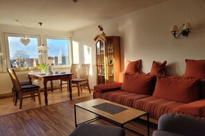 A living room with a sofa, a coffee table, a dining table with chairs, a bookshelf, and a window with a heart decoration. - No. 8 in the Kiebitz House (Borkum)