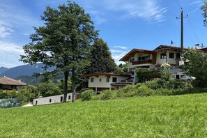 Eine Anhöhe mit mehreren Gebäuden, einem großen Baum und einem Bergpanorama im Hintergrund.