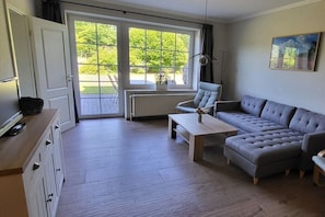 Ein Wohnzimmer mit einer Couch, einem Couchtisch und einem Sessel. Eine Tür führt zu einem Balkon mit Blick auf Grünflächen.