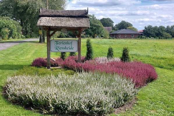 Schild für 'Verienhof Riesenheide' mit einem reetgedeckten Gebäude, umgeben von gepflegten Gärten und einer ländlichen Landschaft.