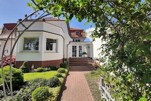 Ein zweistöckiges Gebäude mit weißer Fassade und rotem Dach, ein Hauseingang mit Treppe und ein Garten mit verschiedenen Pflanzen und einem Weg.