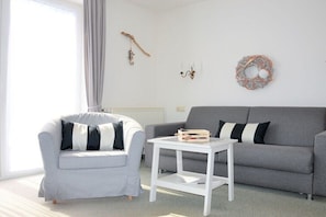 Ein Wohnzimmer mit grauem Sofa, grauem Sessel, weißem Couchtisch und einem Fenster mit grauem Vorhang.