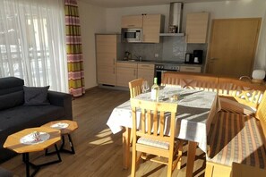 Ein kompakter Wohnbereich mit Sofa, Esstisch und Stühlen, Küchenzeile mit Schränken und Geräten sowie einem Fenster mit Vorhängen.