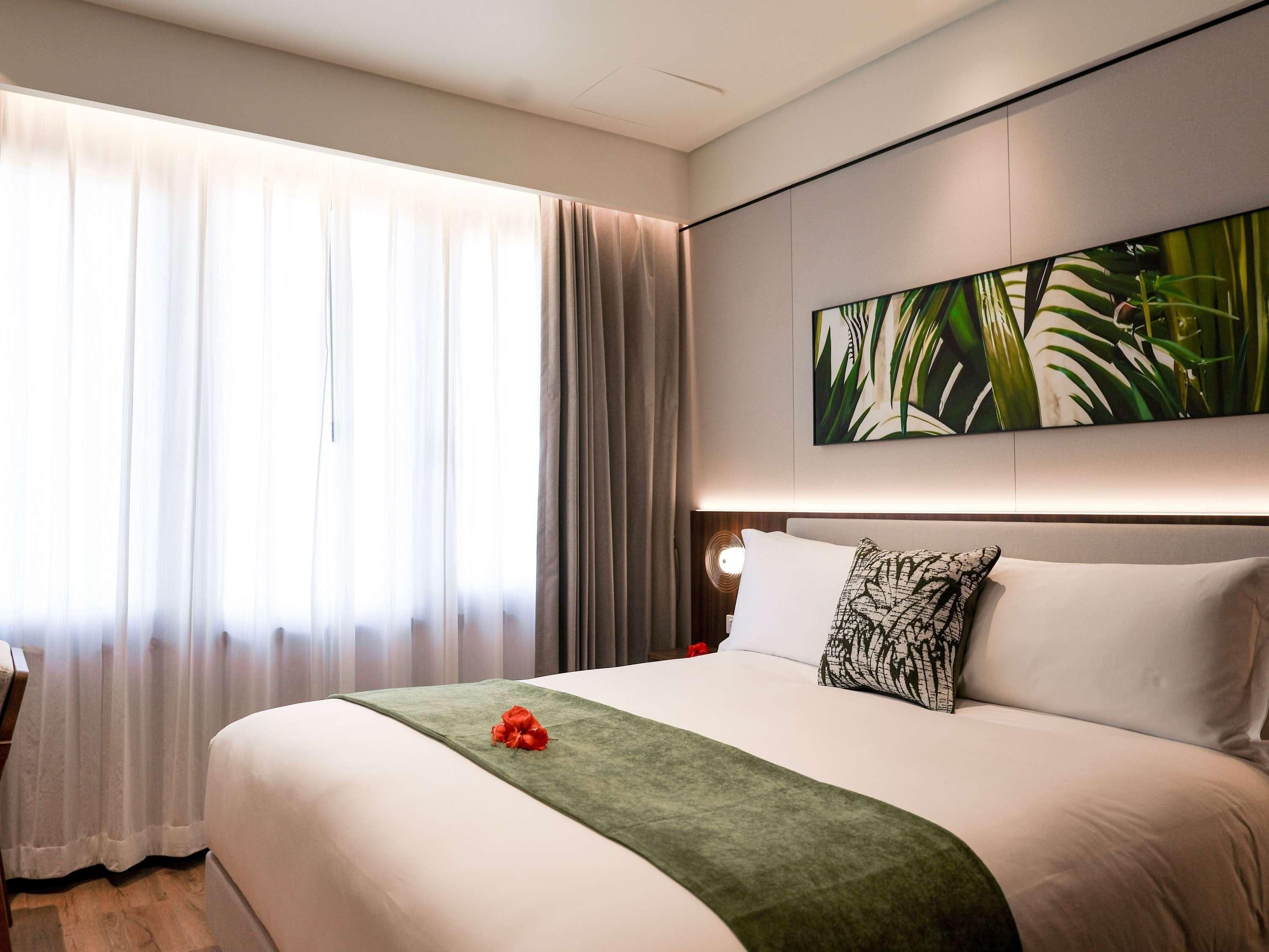 Une chambre d’hôtel avec un grand lit, un plaid vert, un oreiller orné d’un motif de feuilles et une grande fenêtre avec des rideaux légers.