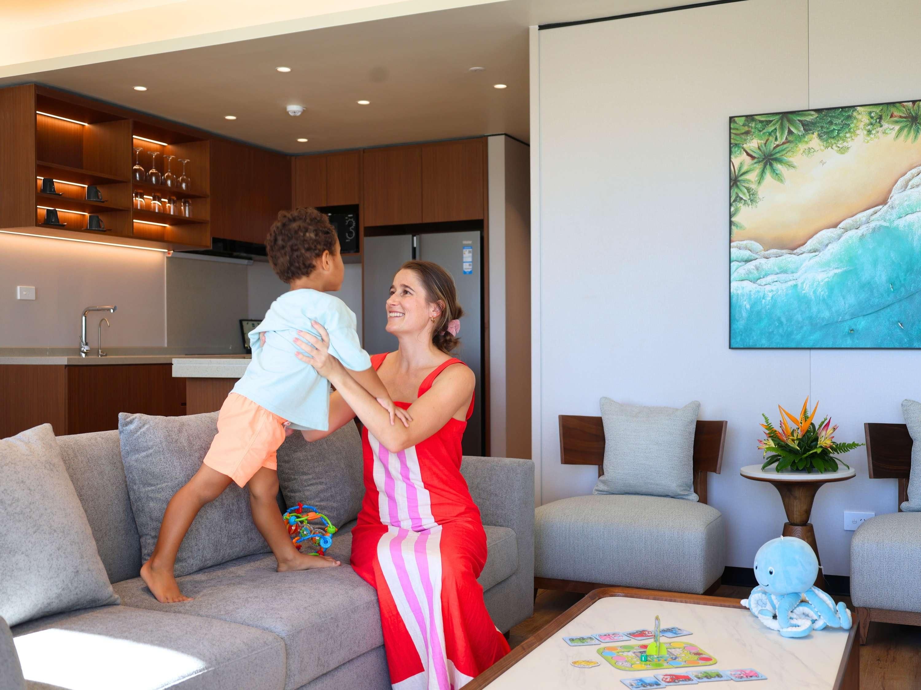 A living room with a sofa, a coffee table with a board game, a child playing, and a kitchen area with cabinets and a refrigerator.