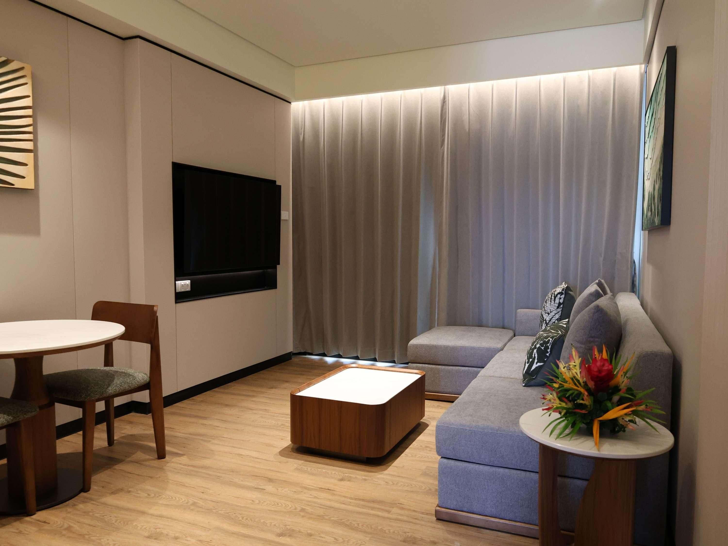 A modern living room with a flat-screen TV, a wooden coffee table, a grey sofa with cushions, and a small round table with a flower arrangement.