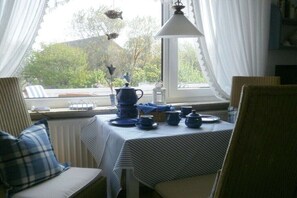Ein Essbereich mit Tisch, der für ein Teebuffet gedeckt ist, Stühlen, einem Fenster mit Vorhängen und Blick auf Grünfläche außerhalb.