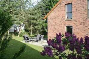 Ein Backsteinhaus mit einer Terrasse, Sitzgelegenheiten im Freien und einem Garten mit violetten Blumen.