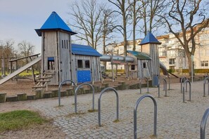 Ein Spielplatz mit Holzspielgeräten und einem blauen Dach, umgeben von einer gepflasterten Fläche und Metall-Bollarden.