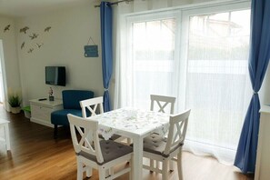 Ein Zimmer mit einem weißen Tisch, vier Stühlen, einem Fernseher auf einem Schrank, einem blauen Sessel, einem Fenster mit Vorhängen und einer Wand mit Vogelaufklebern.