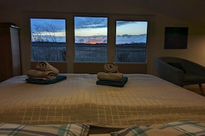 Ein Hotelzimmer mit einem Bett, zwei Fenstern, einem Sessel und einem Gemälde an der Wand.