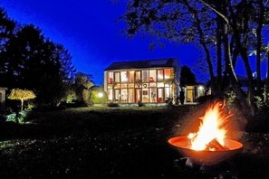Ein modernes Haus mit Glasfassade, einem Feuerkorb im Vordergrund und Bäumen, die das Grundstück umgeben.