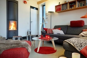 Ein gemütliches Wohnzimmer mit Kamin, einem grauen Sofa mit roten Kissen, einem Glastisch und einem Bücherregal.