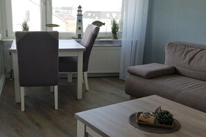 Ein Wohnzimmer mit einer Couch, einem Esstisch mit Stühlen, einem Couchtisch mit Tablett und Blick auf einen Leuchtturm durch ein Fenster.