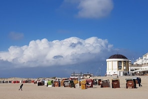 Ein Strand mit einer großen Wolkenformation, einem kleinen Gebäude mit Kuppel und bunten Strandkörben.
