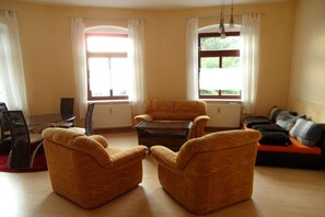Ein Wohnzimmer mit einer Couch, zwei Sesseln, einem Couchtisch, einem Esstisch mit Stühlen und zwei Fenstern mit Vorhängen.