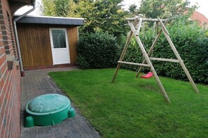 A brick building with a wooden door, a swing set, and a green plastic container. - Holiday home with 1 bedroom (Butjadingen \/ Burhave)