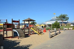 Ein Spielplatz direkt am Strand mit einer Holzkonstruktion, einer überdachten Sitzgelegenheit und einem Gebäude mit einer Flagge im Hintergrund.
