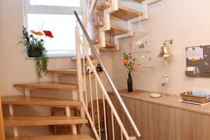 Eine Holztreppe mit Metallgeländer, eine an der Wand befestigte Pflanze, eine dekorative Glocke und ein Schwarzes Brett mit Papieren.