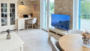 Ein modernes Wohnzimmer mit Schreibtisch, Stuhl, Fernseher und einer Essecke mit Tisch und Stühlen.