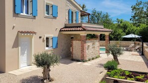 Ein zweistöckiges Haus mit Steinmauern, blauen Fensterläden, einem kleinen Balkon und einem Garten mit Pool und Gartenmöbeln.