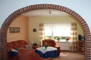 Ein Wohnzimmer mit Backsteinbogen, einer Couch, einem Tisch mit Vase, einem Heizkörper, einem Fernseher und einem Fenster mit Vorhängen.