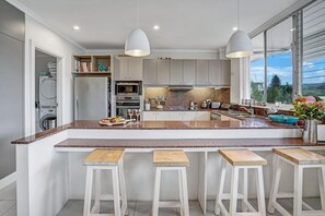 Eine moderne Küche mit Granit Arbeitsplatten, weißen Schränken, einer zentralen Kochinsel mit Barhockern und Blick durch große Fenster.