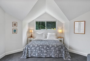 Ein Schlafzimmer mit schrägen Wänden, einem Bett mit gemustertem Überwurf, zwei Nachttischlampen und gerahmten Bildern an den Wänden.