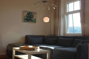 Een woonkamer met een bank, een tafel met dienblad, een schilderij, een lamp en een raam met uitzicht.