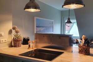 Eine moderne Küche mit einem schwarzen Induktionskochfeld, zwei hängenden Pendelleuchten, einem gewebten Korb mit Küchenutensilien und einem gerahmten Bild an der Wand.