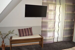 Ein Zimmer mit Bett, Wandfernseher, Fenster mit Vorhängen und einer kleinen Pflanze.