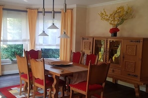 Ein Esszimmer mit einem Holztisch, rot gepolsterten Stühlen, einer Vitrine mit Glastüren und einem Strauß gelbe Blumen an der Wand.
