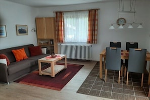 Ein Wohnzimmer mit Sofa, Couchtisch, Esstisch mit Stühlen, einem Heizkörper, Vorhängen, einer Uhr und einem Regal mit Gegenständen.