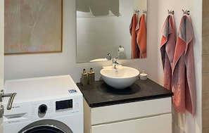 Ein Badezimmer mit einem weißen Waschbecken, einem Spiegel, einer Waschmaschine und Handtüchern auf einem Ablagegestell.
