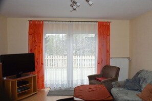 Ein Wohnzimmer mit einem Fernseher auf einem Holzboden, einem Sofa, einem Sessel und einem Fenster mit Vorhängen.