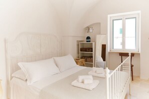 Ein Schlafzimmer mit einem weißen Bett, einem Holfnachtkästchen, einem Regal mit Körben und einem Fenster mit Jalousien.