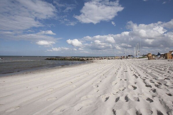 Ein sandiger Strand mit Fußabdrücken, eine felsige Küstenlinie und Segelboote in der Ferne.